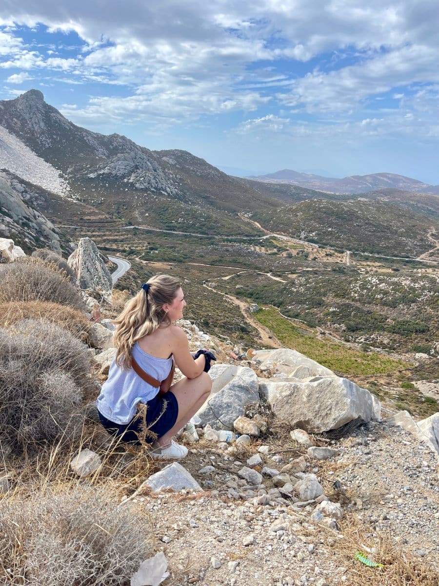 A woman crouches on rocky terrain, gazing over a vast, rugged landscape under a partly cloudy sky. She appears to be in a remote, mountainous area, surrounded by dry brush and boulders, enjoying a peaceful, contemplative moment.