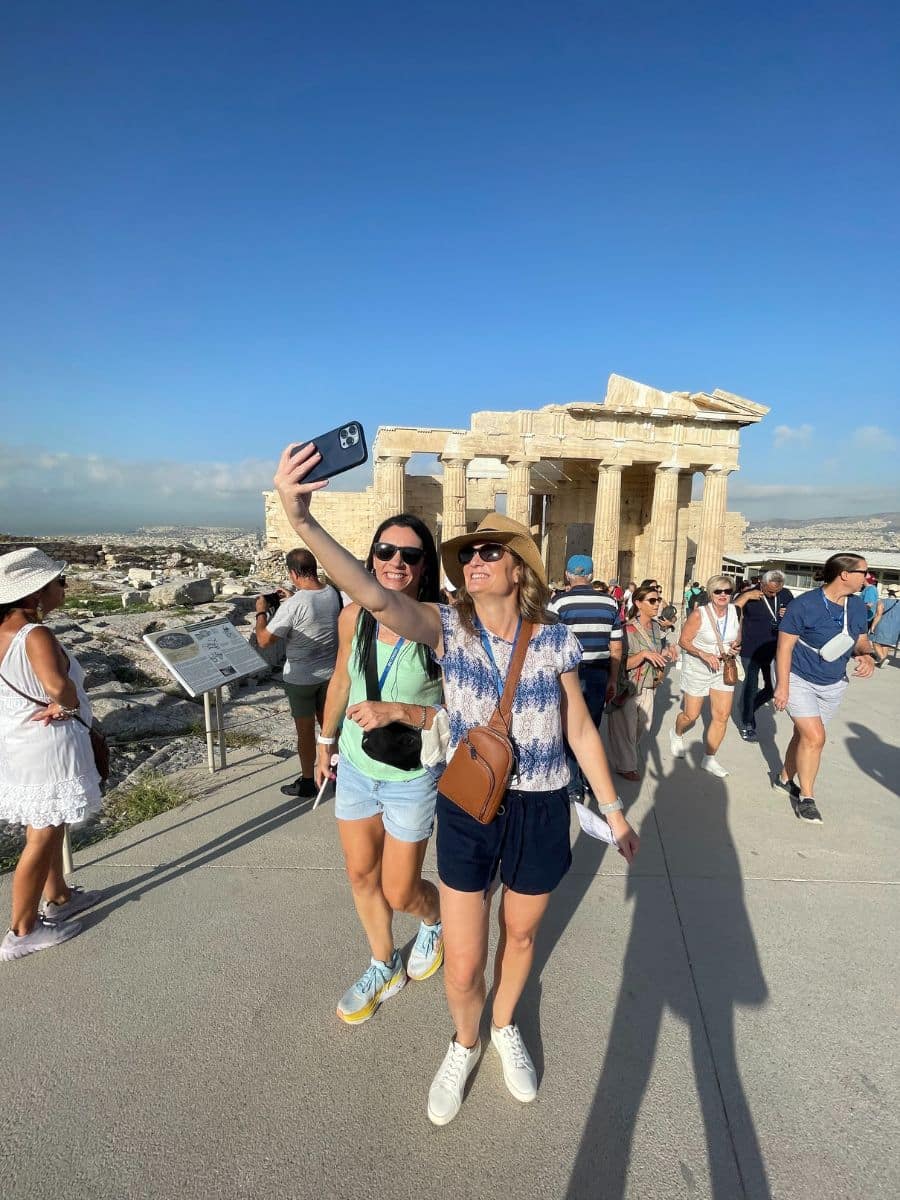 Two women taking a selfie in front of ancient ruins, possibly the Acropolis in Greece. They smile as they capture the moment, with other tourists and bright blue skies in the background, embodying the lively experience of traveling with friends.