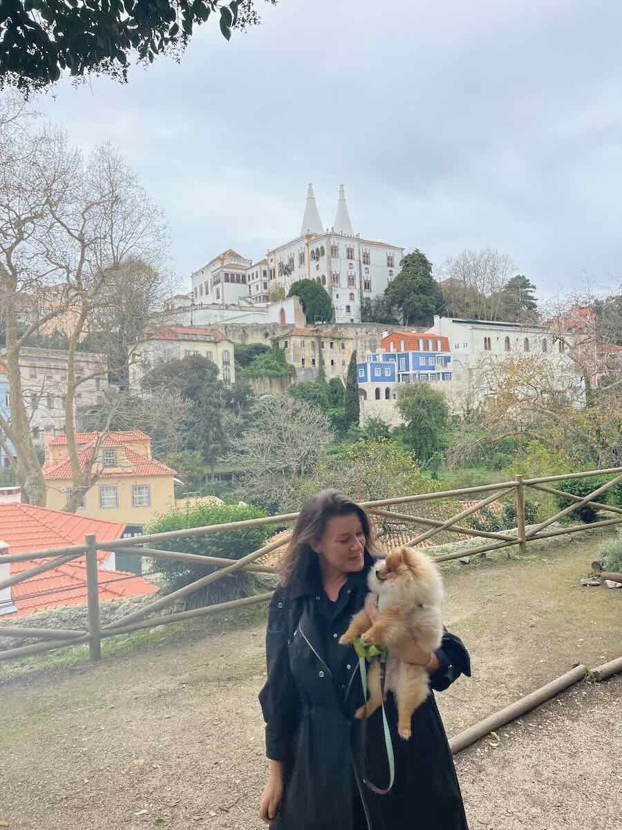 Melissa with Teddy, stands in front of a scenic hillside view featuring colorful buildings and the iconic spires of the National Palace of Sintra in Portugal.