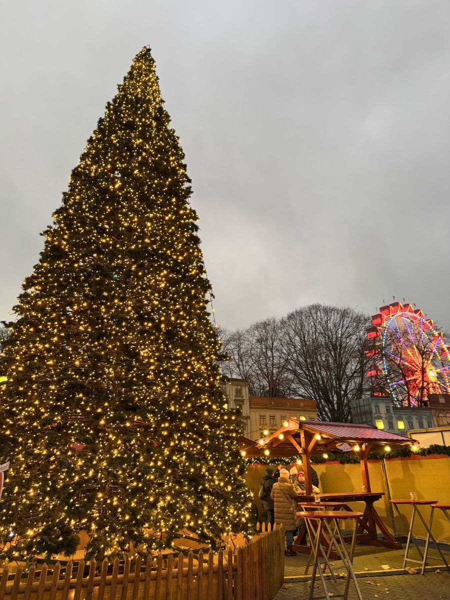 A giant Christmas tree covered in golden fairy lights stands tall, surrounded by wooden stalls and tables. In the background, a brightly lit Ferris wheel adds to the festive atmosphere, with people mingling at the market.