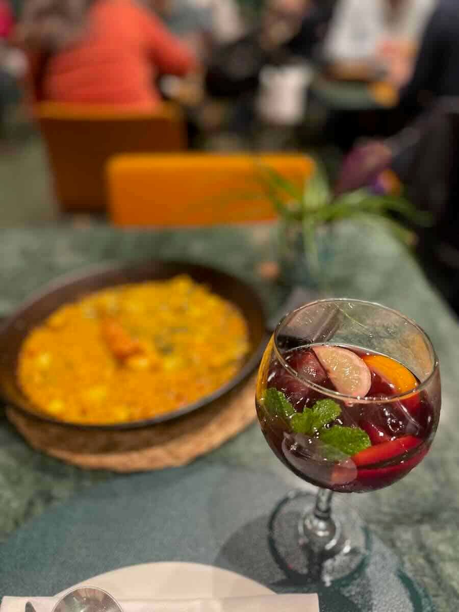 A glass of sangria garnished with fresh fruit sits in the foreground, with a paella dish blurred in the background, capturing a quintessential solo dining experience in Valencia.