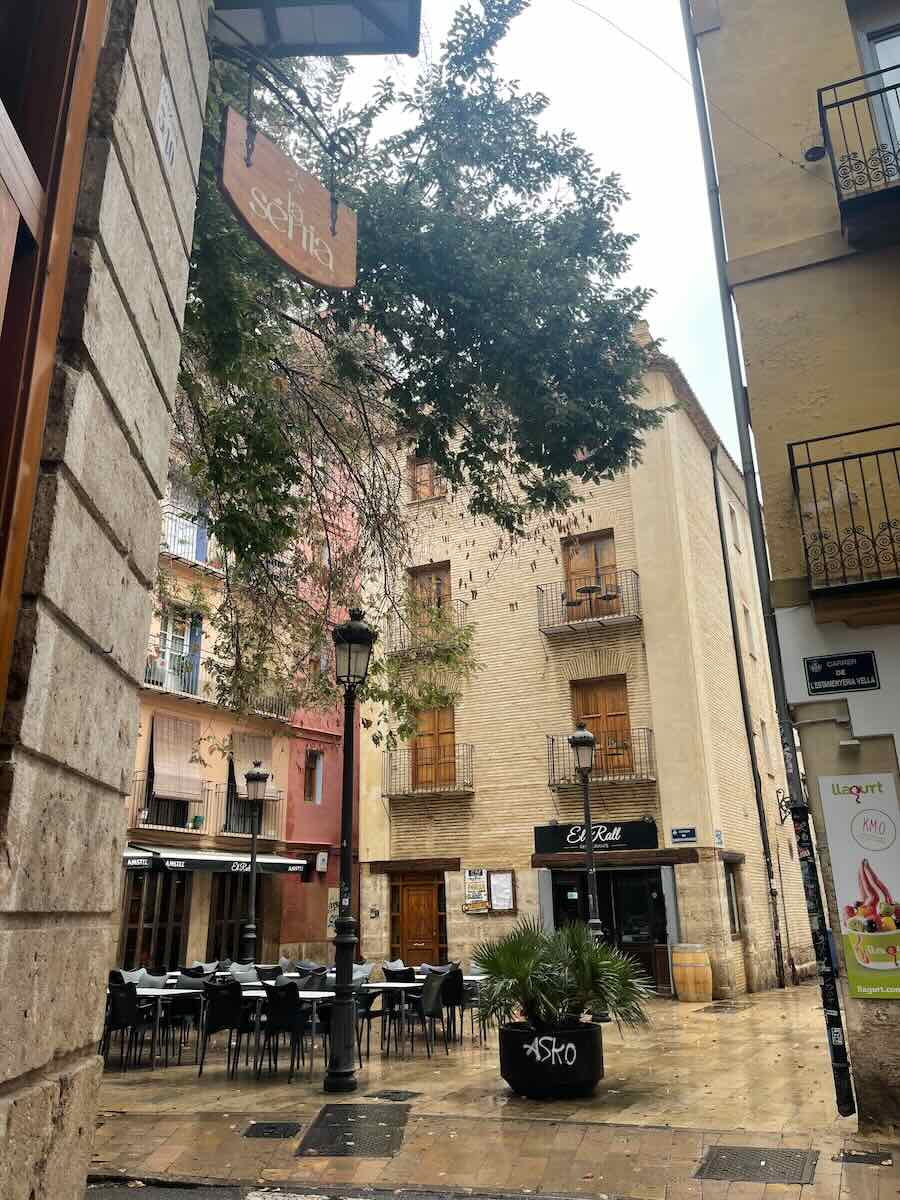 A charming street in Valencia lined with colorful buildings and outdoor seating. The sign for "La Senia" hangs nearby, with a few trees and traditional street lamps enhancing the picturesque atmosphere.