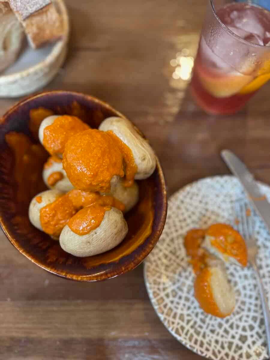 A bowl of Valencia-style patatas bravas topped with a rich, red sauce, alongside a drink and basket of bread, perfect for a solo traveler indulging in local tapas.