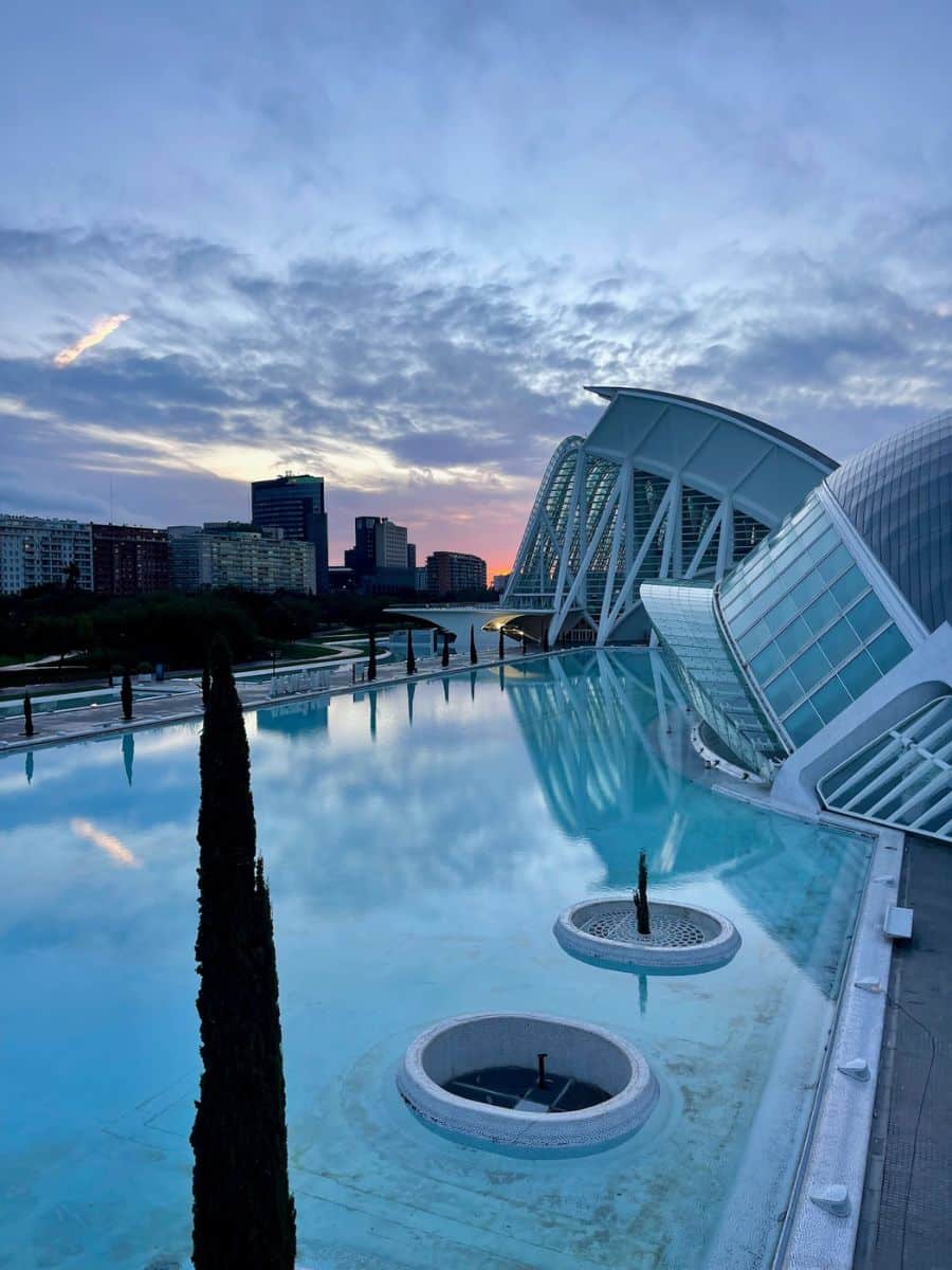Reflections on the still water in front of modern architecture at the City of Arts and Sciences in Valencia, Spain, during twilight. The sky is partly cloudy with a hint of the setting sun in the distance. Sleek, curved buildings with glass and white structural elements are mirrored in the water, adding to the serene atmosphere.