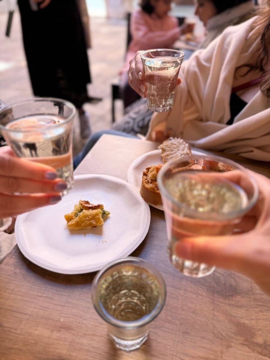 A cozy moment at a Venetian café as friends toast with glasses of white wine over a table with small bites and pastries, capturing the warmth and joy of shared travel experiences.