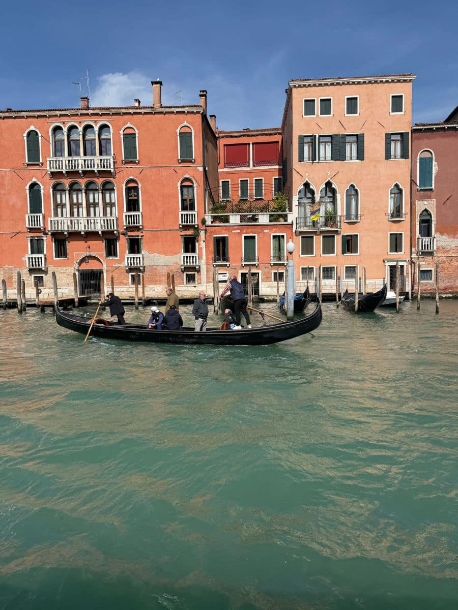 A picturesque view of the Grand Canal in Venice, featuring a gondola with several passengers enjoying a ride. The gondolier stands at the stern, using his oar to steer the boat. Historic buildings in shades of orange and beige with arched windows and balconies overlook the waterway.