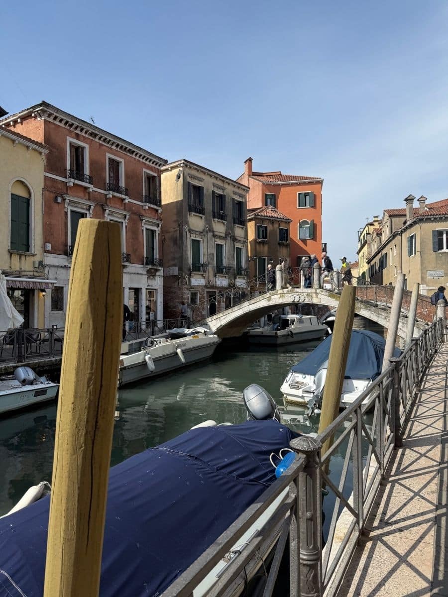 A picturesque scene in Venice featuring a stone pedestrian bridge spanning a canal filled with boats. Buildings in various shades of orange and yellow with multiple windows overlook the canal. The view is taken from a walkway alongside the water, with wooden mooring posts in the foreground.