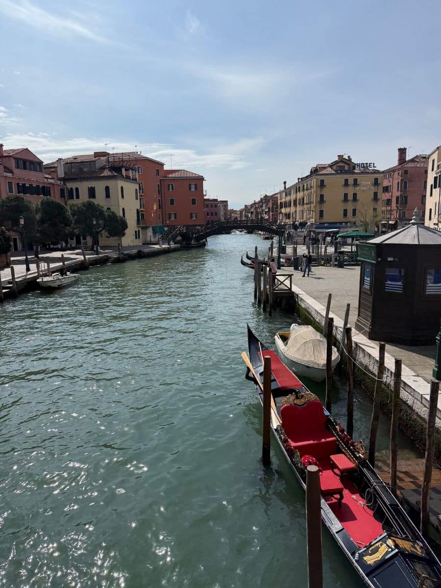 A wide view of a bustling canal in Venice, Italy, with colorful buildings lining both sides. Several gondolas and other boats are moored along wooden pilings. A small kiosk or booth stands on the right bank, and people can be seen walking along the waterfront. The water reflects the buildings and the partly cloudy sky.