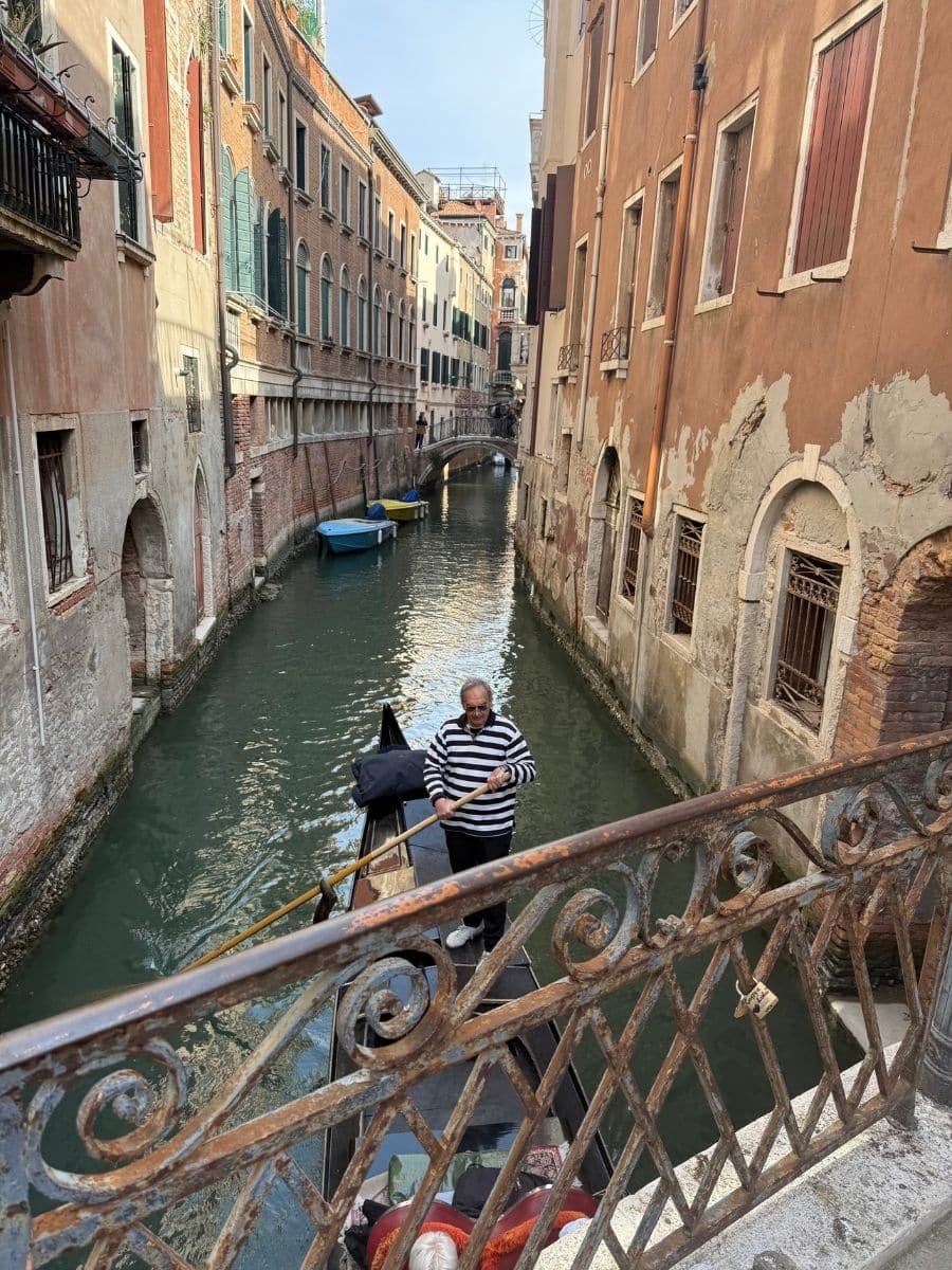 A gondolier in a black and white striped shirt propels a gondola down a narrow canal in Venice, Italy. Tall, colorful buildings with closed shutters line both sides of the waterway. The view is captured from a bridge with ornate metal railings.