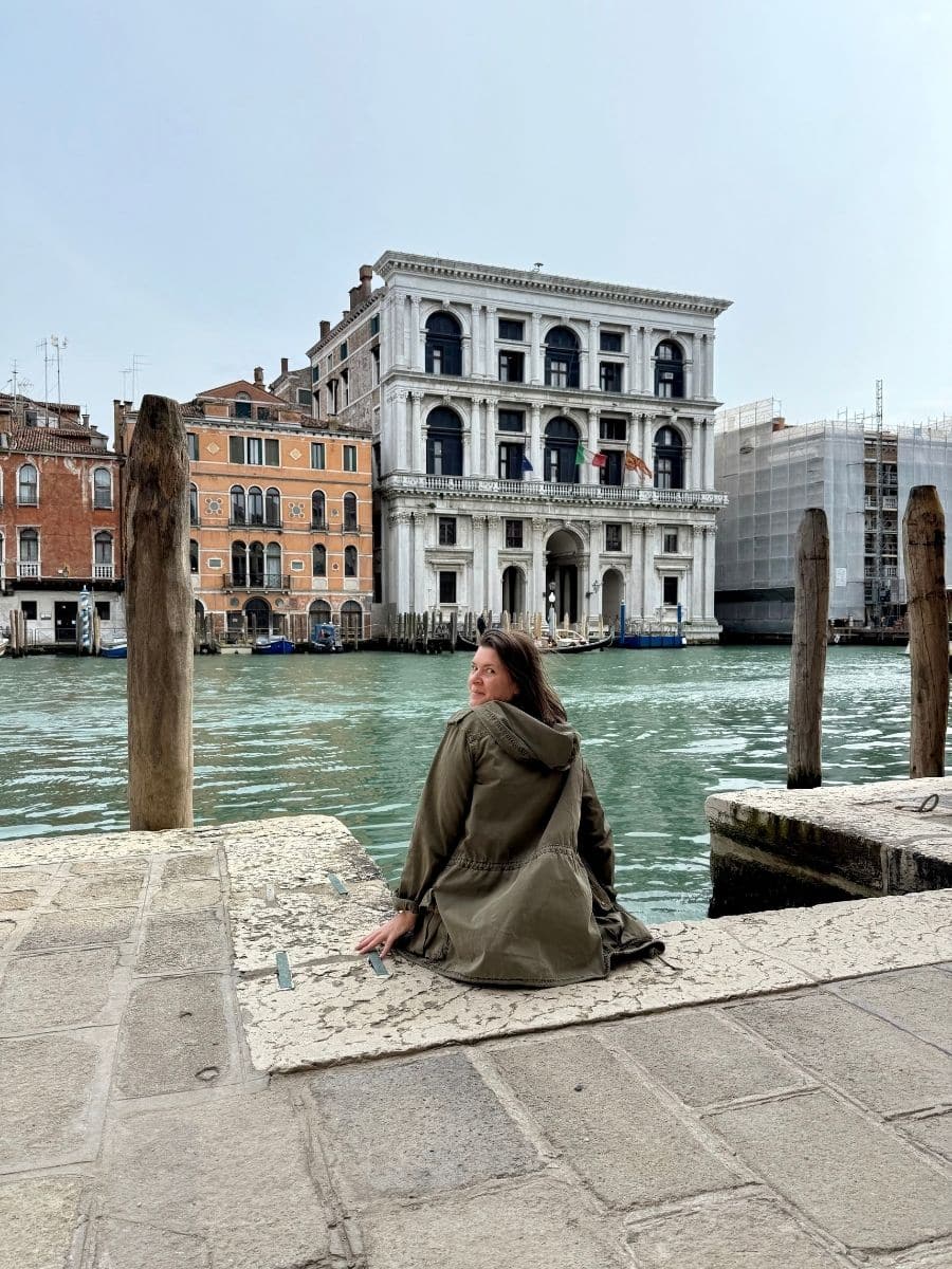 Melissa wearing a green jacket sits on the stone edge of the Grand Canal in Venice, Italy, looking over her shoulder at the camera. In the background, the Fondaco dei Turchi, a large white palazzo with arched windows and flags, stands prominently along the waterway. Other colorful Venetian buildings line the canal under an overcast sky.