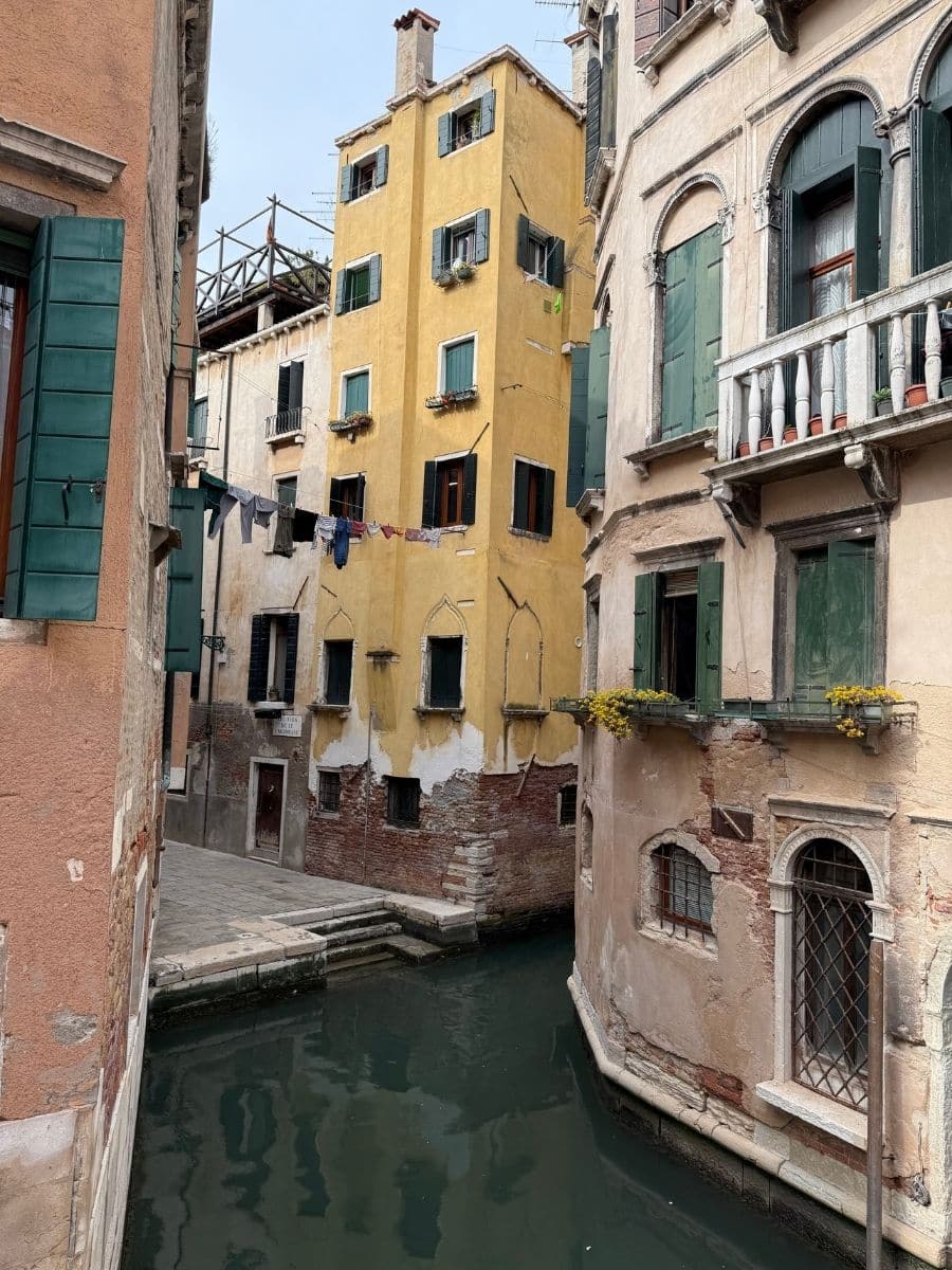 A canal intersection in Venice, Italy, where a narrow waterway meets a slightly wider one. Colorful, aged buildings with green shutters line both sides of the canals. A stone fondamenta (walkway) runs along the edge. Laundry hangs between buildings, adding a touch of everyday Venetian life.