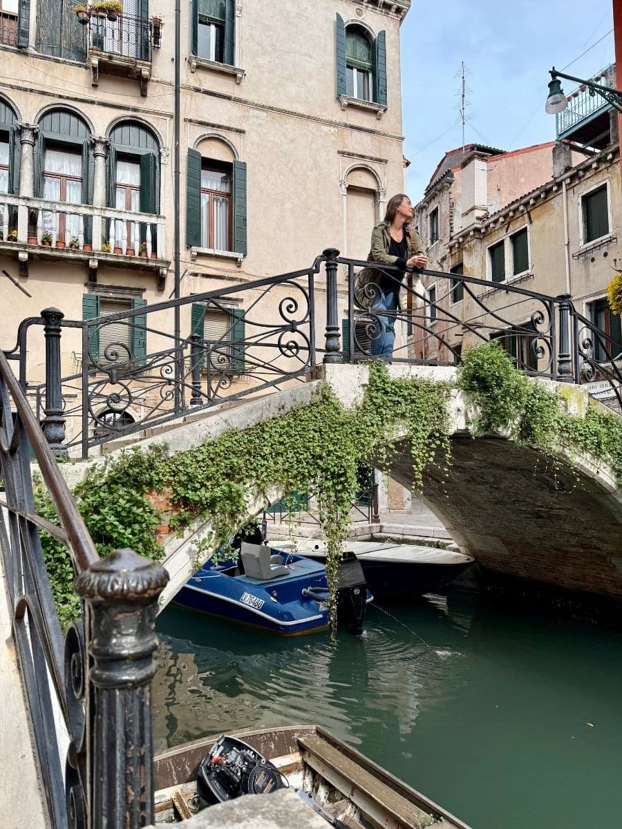 Melissa wearing a patterned jacket and jeans stands on a small, arched pedestrian bridge covered in ivy, looking down at the green canal below in Venice, Italy. Colorful, aged buildings with window shutters line the waterway. A small blue boat is moored beneath the bridge.