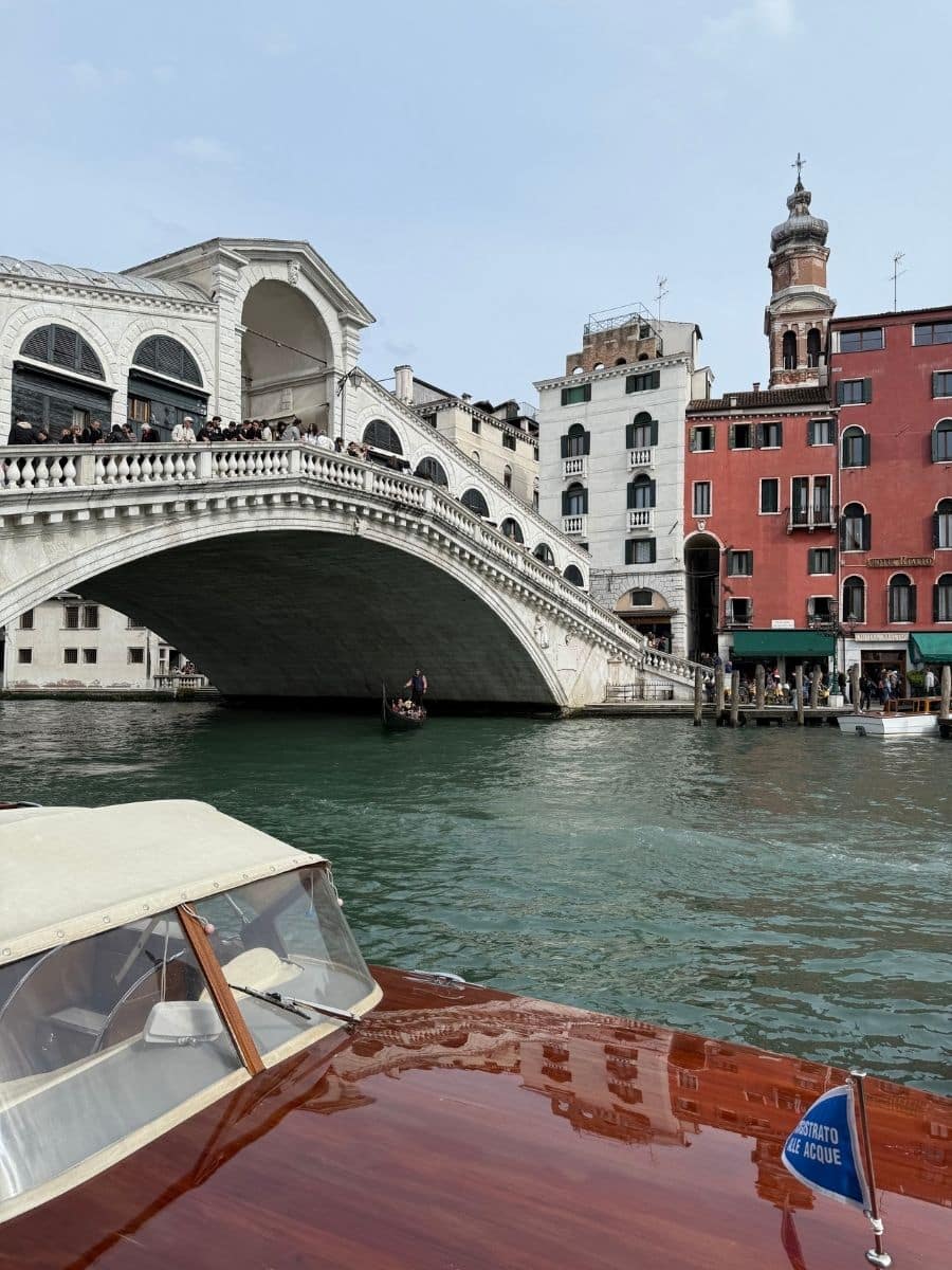 The grandeur of the Rialto Bridge as it arches over the bustling Grand Canal in Venice. Tourists stroll along the bridge, taking in the sights. Historic buildings in various shades of red, white, and beige create a vibrant backdrop. The foreground shows the polished wood and windshield of a boat navigating the canal, with a small flag indicating "STRATO ALE ACQUE."
