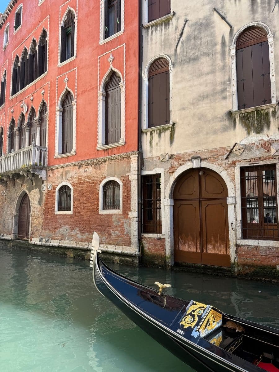 A gondola floats peacefully on a narrow canal in Venice, Italy, beside colorful, historic buildings with ornate arched windows and shuttered doors.