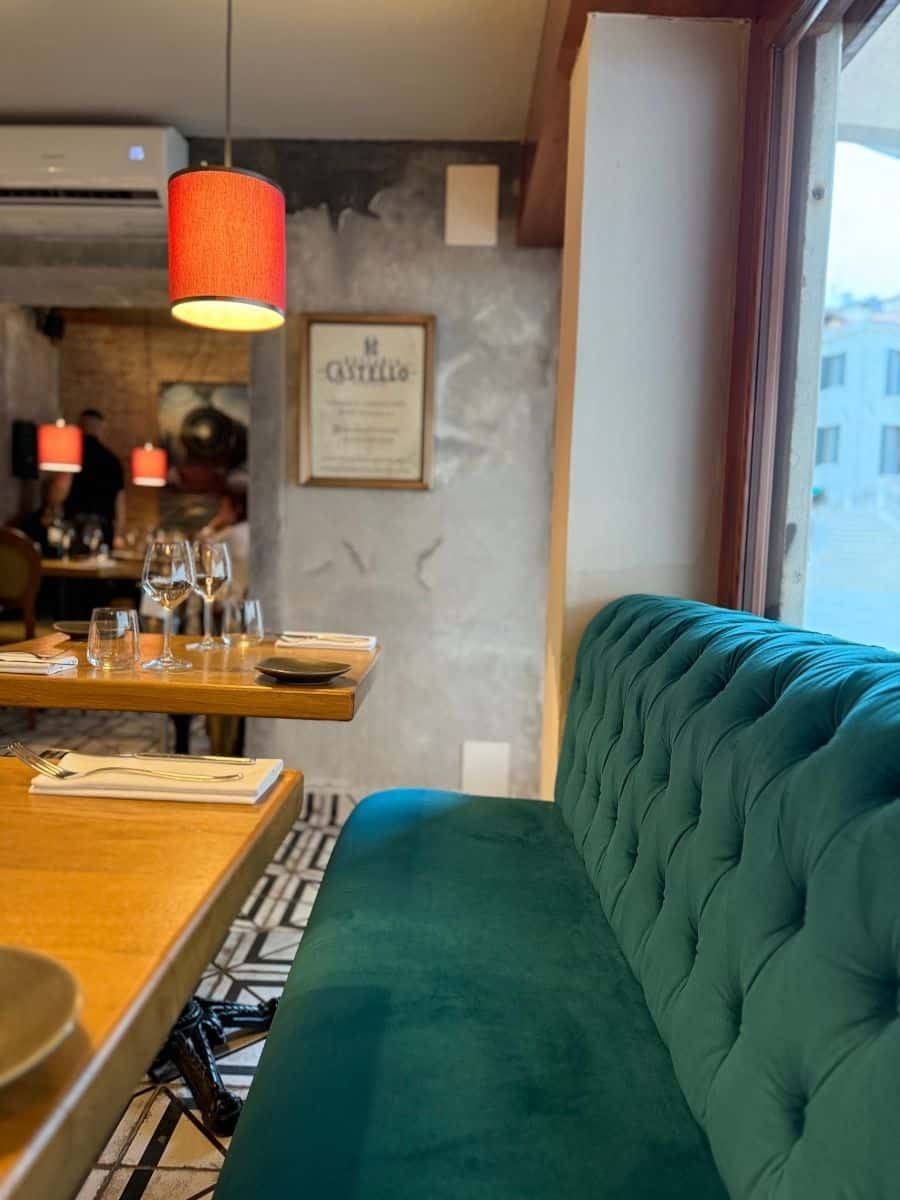 The interior of a restaurant features a plush, tufted teal velvet banquette in the foreground, extending along a window that offers a glimpse of the outside. Several wooden tables with place settings, including wine glasses and cutlery, are arranged in the dining area. The lighting is warm, with a red pendant lamp hanging above one table. A framed certificate or artwork is visible on the textured gray wall.