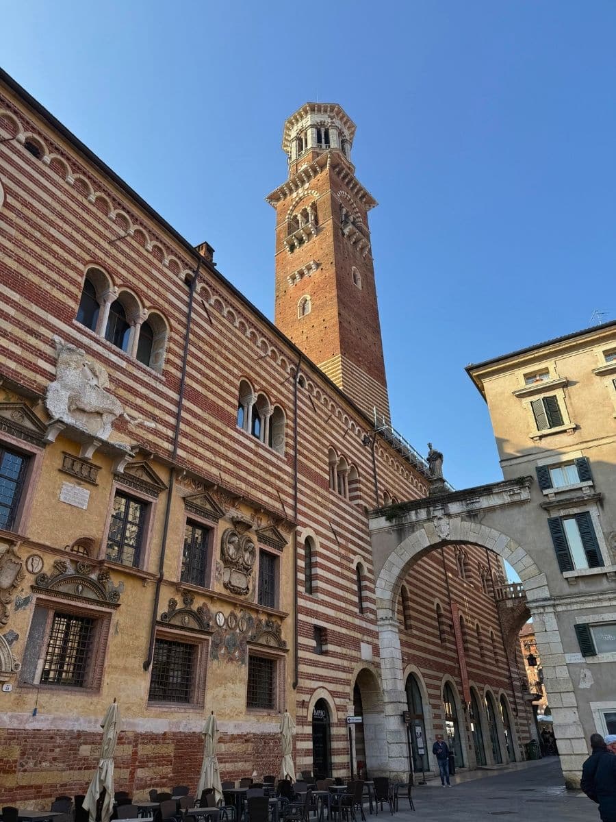 A striking view of Verona’s Torre dei Lamberti rising above a richly decorated medieval building with alternating red brick and white stone stripes. The scene includes an arched walkway and a small piazza with café tables below, all under a clear blue sky.