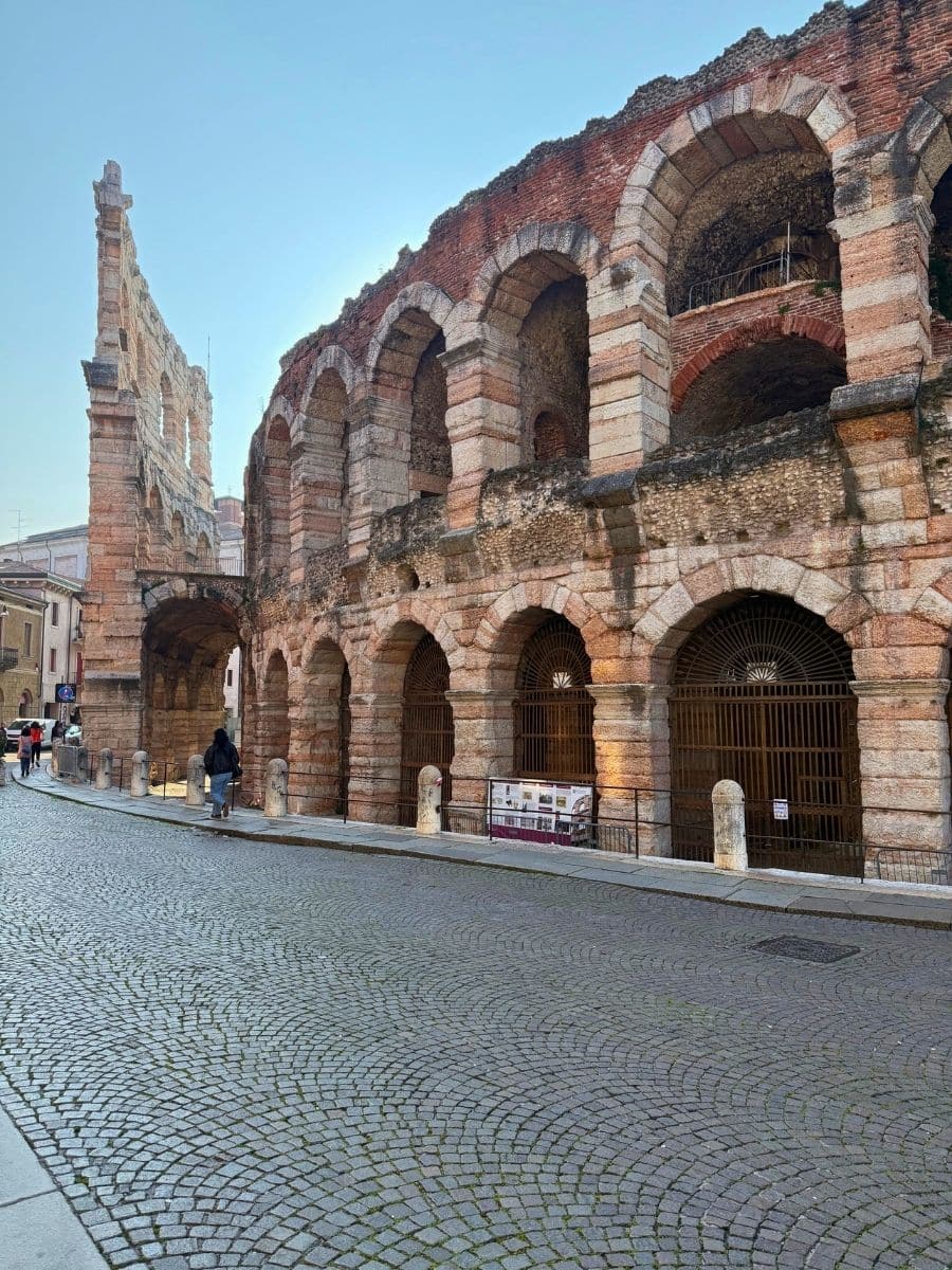 Ancient Roman amphitheater in Verona, Italy, known as the Verona Arena, featuring large arched stone entrances and weathered pink limestone walls. The cobblestone street in front is quiet with a few pedestrians, and the early morning or late afternoon light casts soft shadows across the scene.