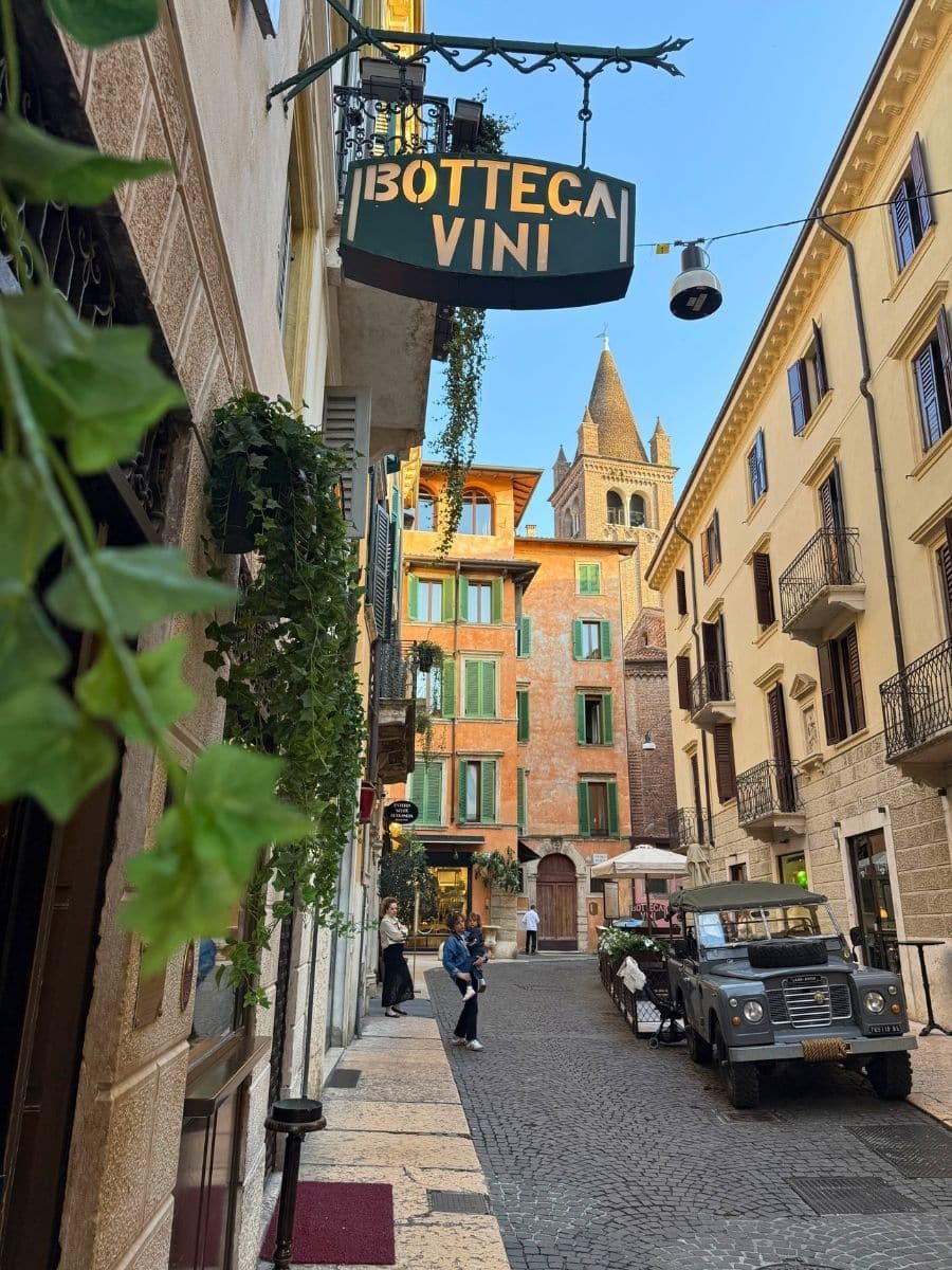 A charming narrow street in Verona, Italy, featuring a hanging sign for “Bottega Vini,” classic architecture with balconies and green shutters, a vintage Land Rover parked near a small outdoor seating area, and a few people strolling along the cobblestone path under a clear blue sky.