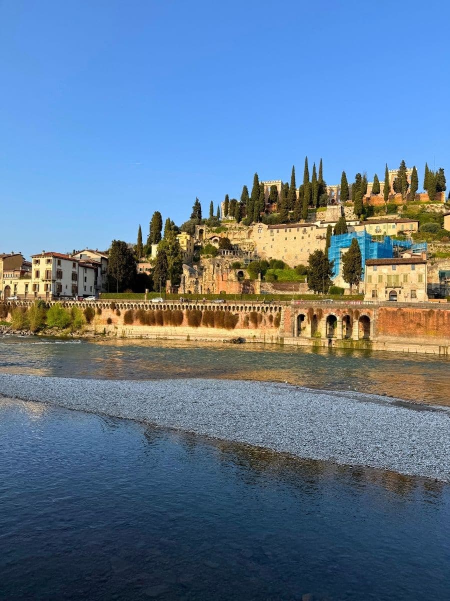 A scenic view of Verona, Italy, featuring the Adige River in the foreground with clear, rippling water. In the background, a hillside is dotted with tall cypress trees and historic stone buildings, including Roman ruins and structures covered partly in scaffolding, all bathed in warm afternoon sunlight under a bright blue sky.