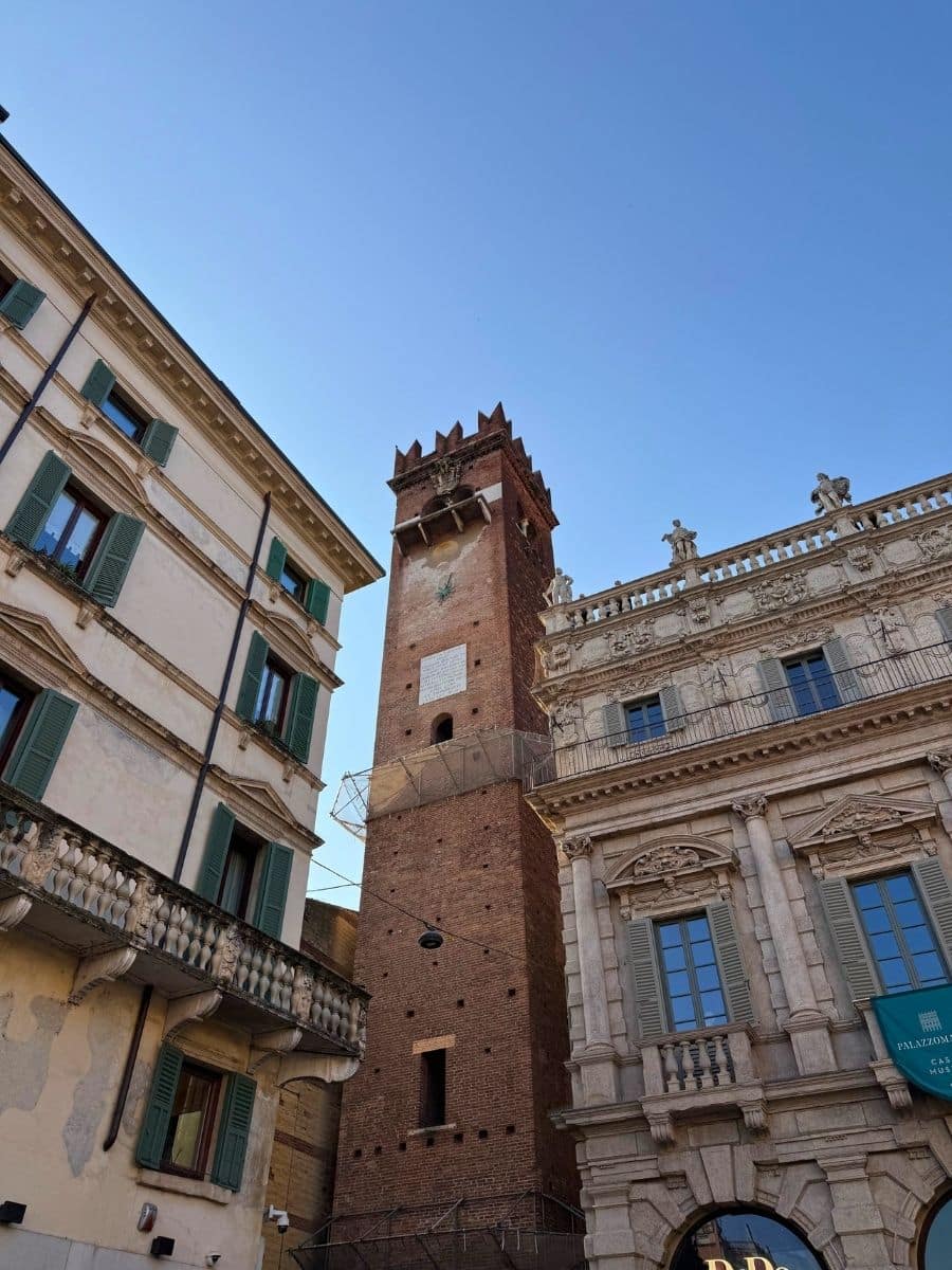 An upward angle captures the Torre dei Lamberti in Verona, Italy, a tall brick tower with a crenellated top, standing between two ornate buildings under a clear blue sky. The building on the left is cream-colored with green shutters and decorative balconies, while the building on the right is more detailed with arched windows and stone carvings.