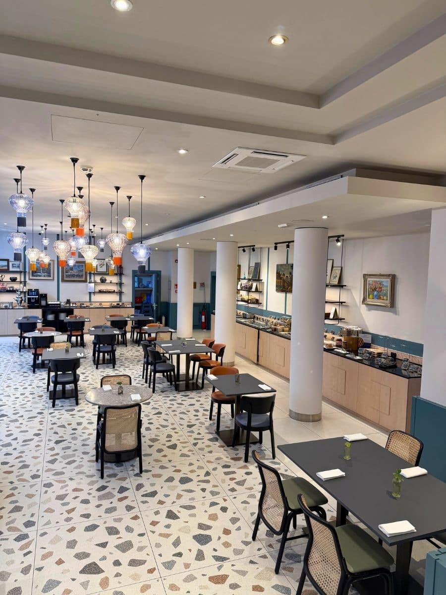 A brightly lit and spacious dining area, likely a hotel restaurant in Verona, featuring numerous tables and chairs arranged on a patterned terrazzo floor. A long buffet area with various food selections lines the back wall, while modern pendant lights with colorful glass shades hang from the ceiling.