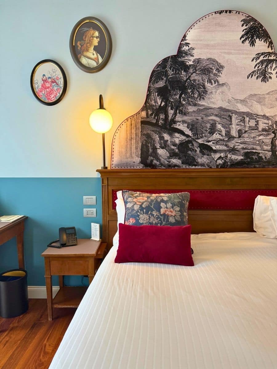 A stylish hotel room, likely at the Hotel Indigo Verona, featuring a bed with white linens, a floral patterned pillow, and a deep red lumbar pillow. A wooden headboard with a decorative mural of a landscape extends upwards, and two round framed pictures hang on the light blue wall above a modern wall lamp. A small wooden nightstand holds a retro-style telephone and a small booklet.