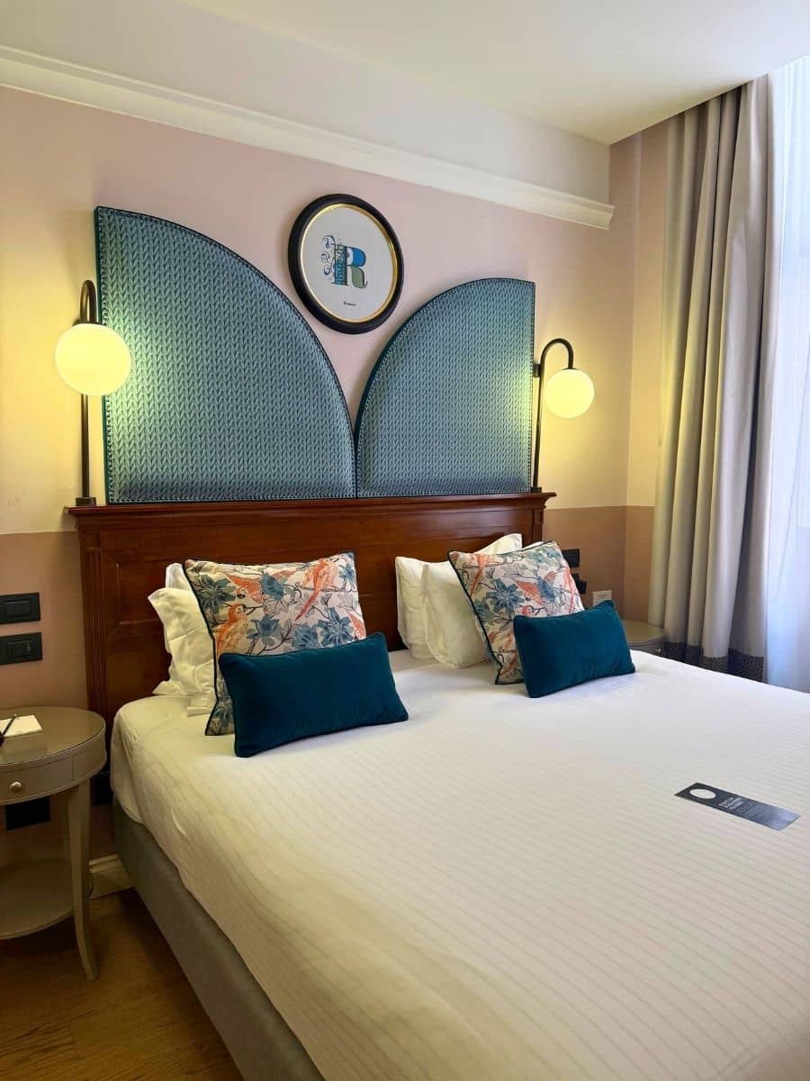 A well-appointed hotel room, likely in Verona, featuring a bed with crisp white linens, floral patterned pillows, and teal lumbar pillows. A wooden headboard with teal patterned panels is flanked by modern wall lamps with round shades. Above the bed, a round framed picture with a stylized "R" hangs on the light pink wall. A small round nightstand is visible on the left side of the bed.