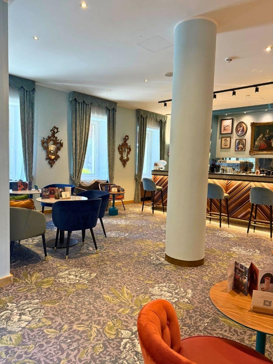 A stylish lounge area, likely within the Hotel Indigo Verona, featuring a mix of seating arrangements on a patterned carpet. Several small round tables with plush chairs in shades of blue and orange are scattered throughout the space, while a sleek bar area with blue stools and warm lighting is visible in the background, adorned with framed artwork on the wall. Large windows with patterned curtains let in natural light, contributing to the inviting and modern atmosphere.