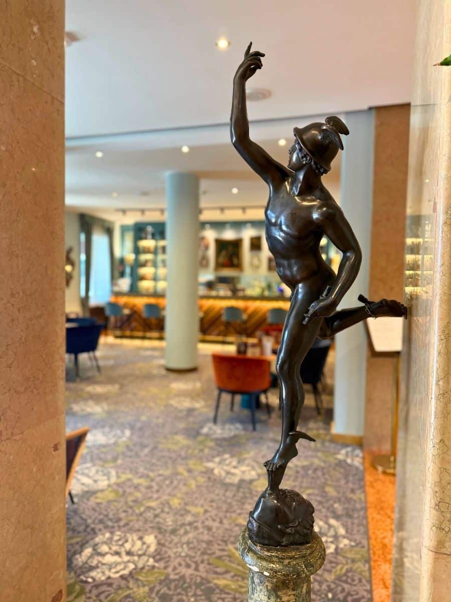 This interior shot, likely within the Hotel Indigo Verona, focuses on a bronze statue of a winged figure wearing a helmet, posed dynamically with one arm raised and one leg extended. The statue rests on a decorative marble pedestal. The blurred background reveals a stylish lounge or bar area with patterned carpets, plush seating in shades of blue and orange, and a well-lit bar with wooden accents and framed artwork on the walls. The overall ambiance suggests a sophisticated and artistic space.