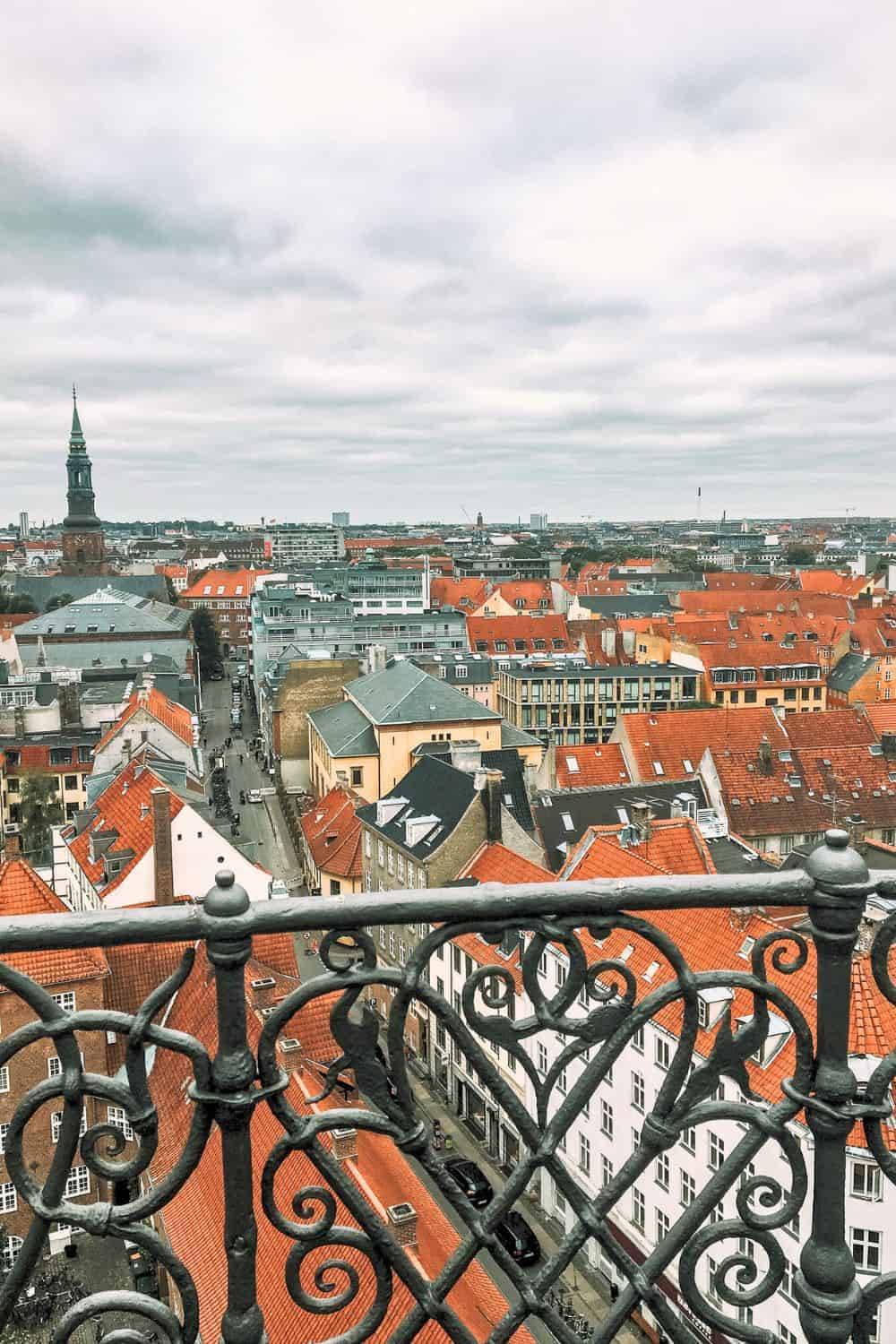 Panoramic view of Copenhagen's cityscape featuring a sea of red rooftops, viewed from an ornate balcony, with the spire of St. Peter's Church in the distance.