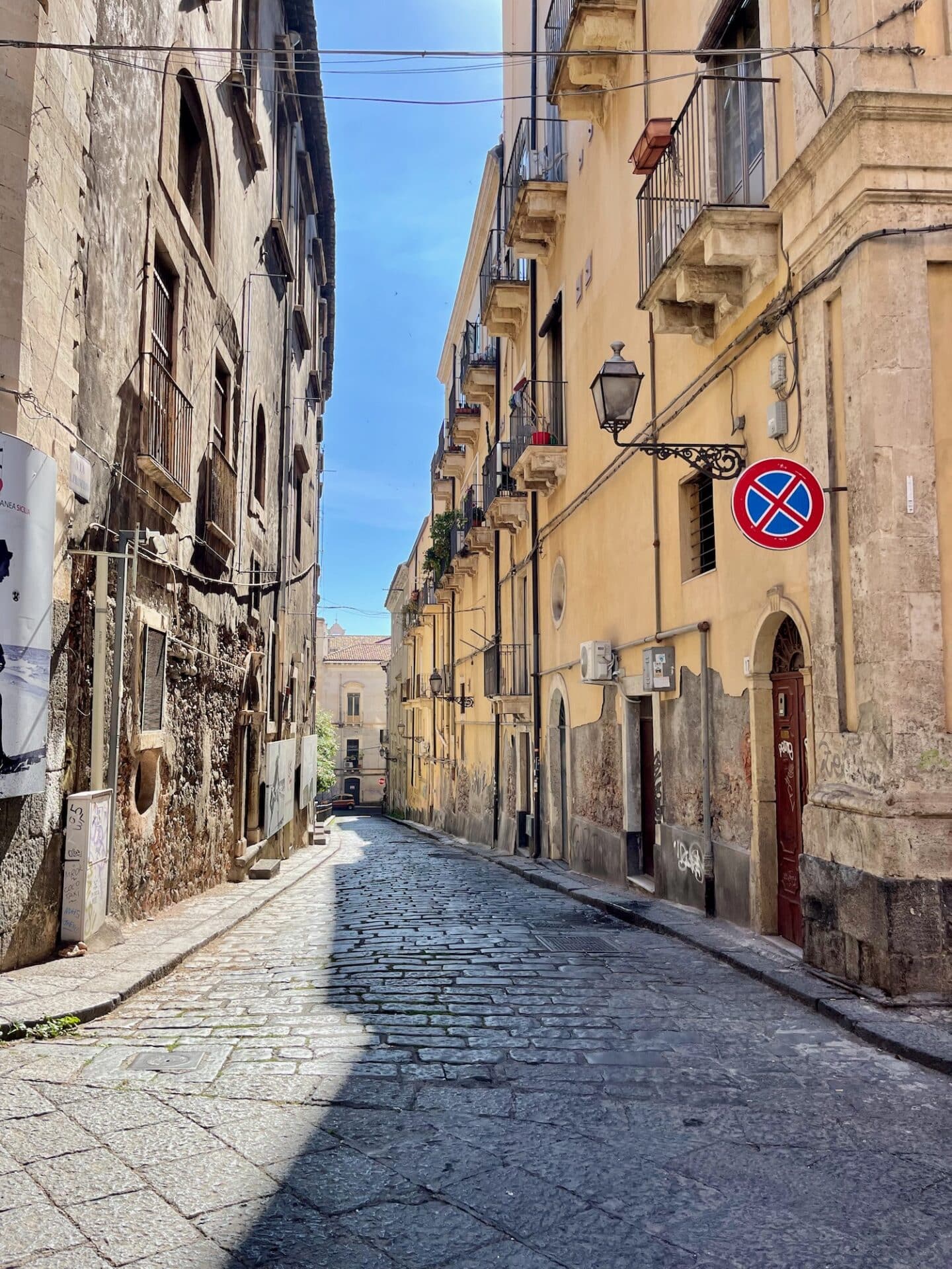 Narrow cobblestone street in Catania flanked by old buildings with balconies and a "no parking" sign.