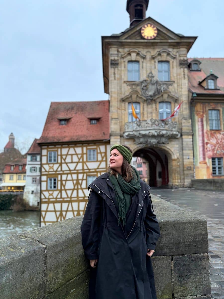 a solo female traveler stands on a bridge in Europe, dressed warmly for winter. Behind her is a charming, historic town hall, illustrating the scenic and rich history that European cities offer to solo explorers.