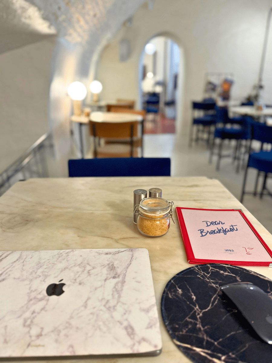 A marble-topped table at 'Dear Breakfast Bica,' featuring a marble-patterned laptop, a red menu, a black marble mouse pad, and a jar of brown sugar. The cozy café interior with arched doorways is visible in the background.