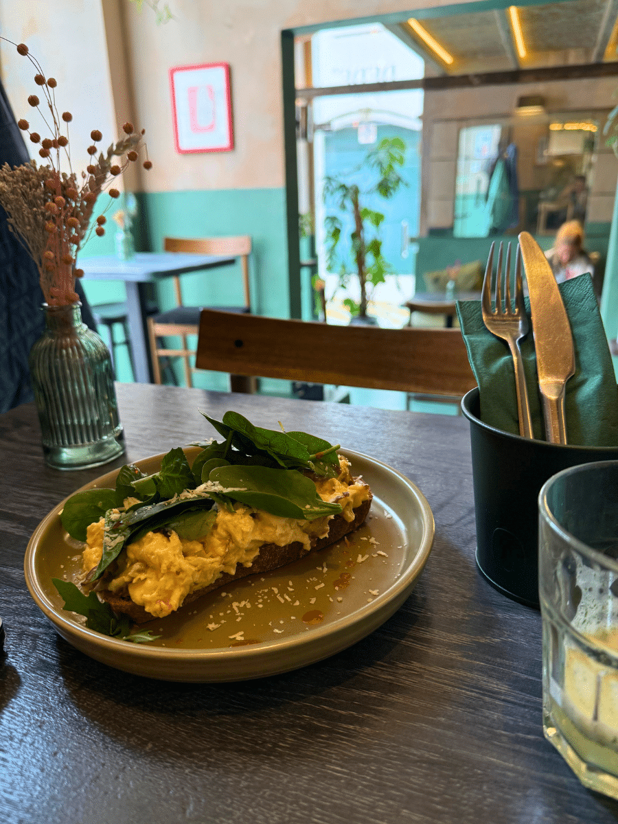 A plate of avocado toast topped with scrambled eggs and greens at 'Dede's Cafe.' The table includes a vase with dried flowers and cutlery in a green napkin holder. The cozy cafe interior with green accents and large windows is visible in the background.