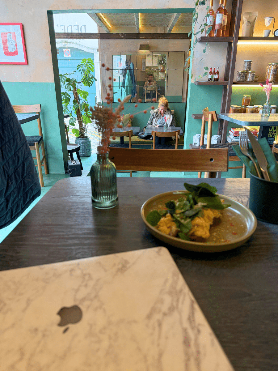 Inside Dede's Cafe, a cozy setting with green and beige walls, wooden furniture, and potted plants. A laptop with a marble-patterned cover sits on a table next to a plate of scrambled eggs on toast. In the background, a woman sits at a table reading, with a mirror reflecting the room's warm ambiance.