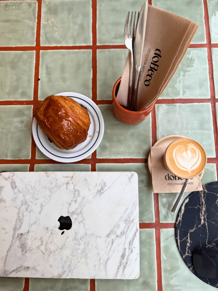 A flat-lay shot on a green-tiled table at "Do Beco" café, showcasing a marble-textured laptop, a golden pain au chocolat, a cup of latte art, and utensils wrapped in branded napkins with "Do Beco" written on them.