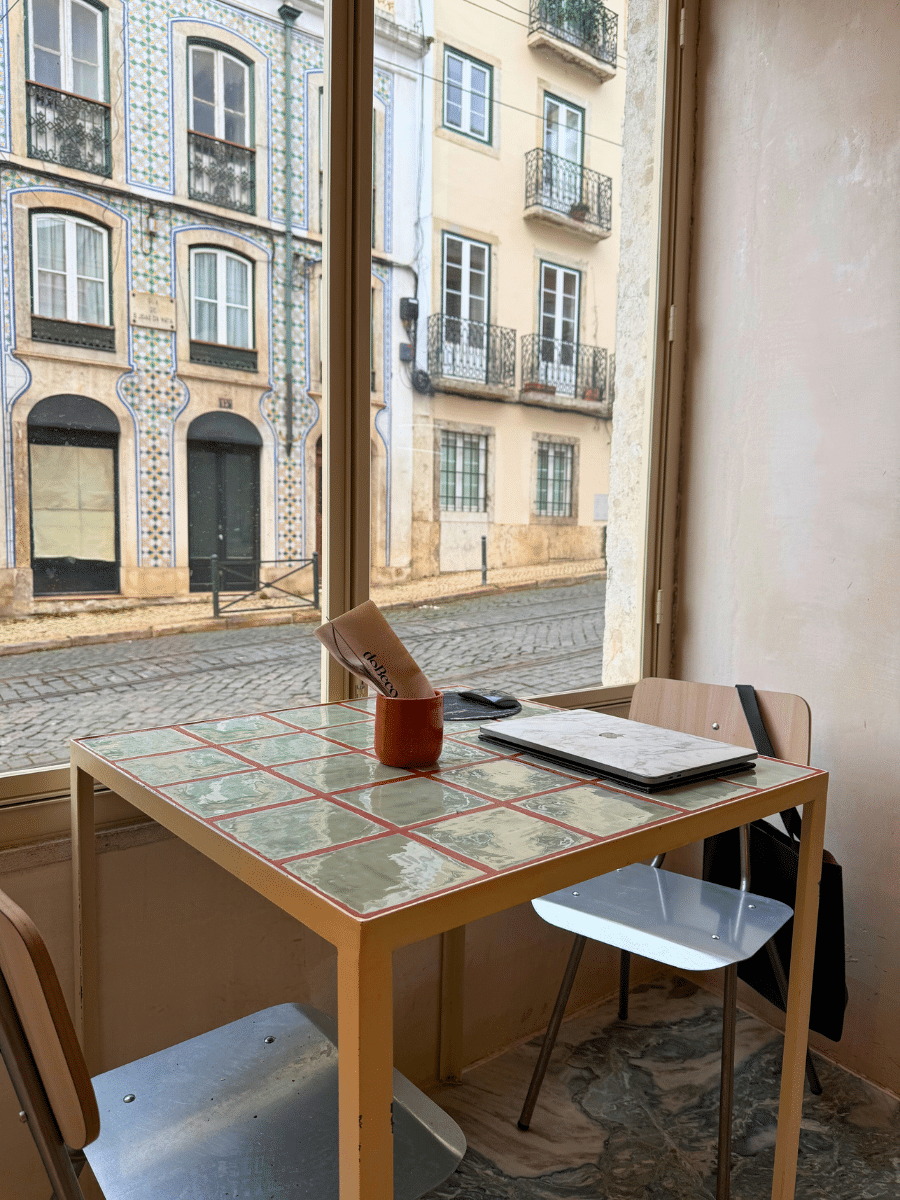 A cozy table at "Do Beco" café, featuring a marble-textured laptop, utensils in a terracotta holder with "Do Beco" branding, and a window view of traditional Portuguese tiled buildings and cobblestone streets outside.