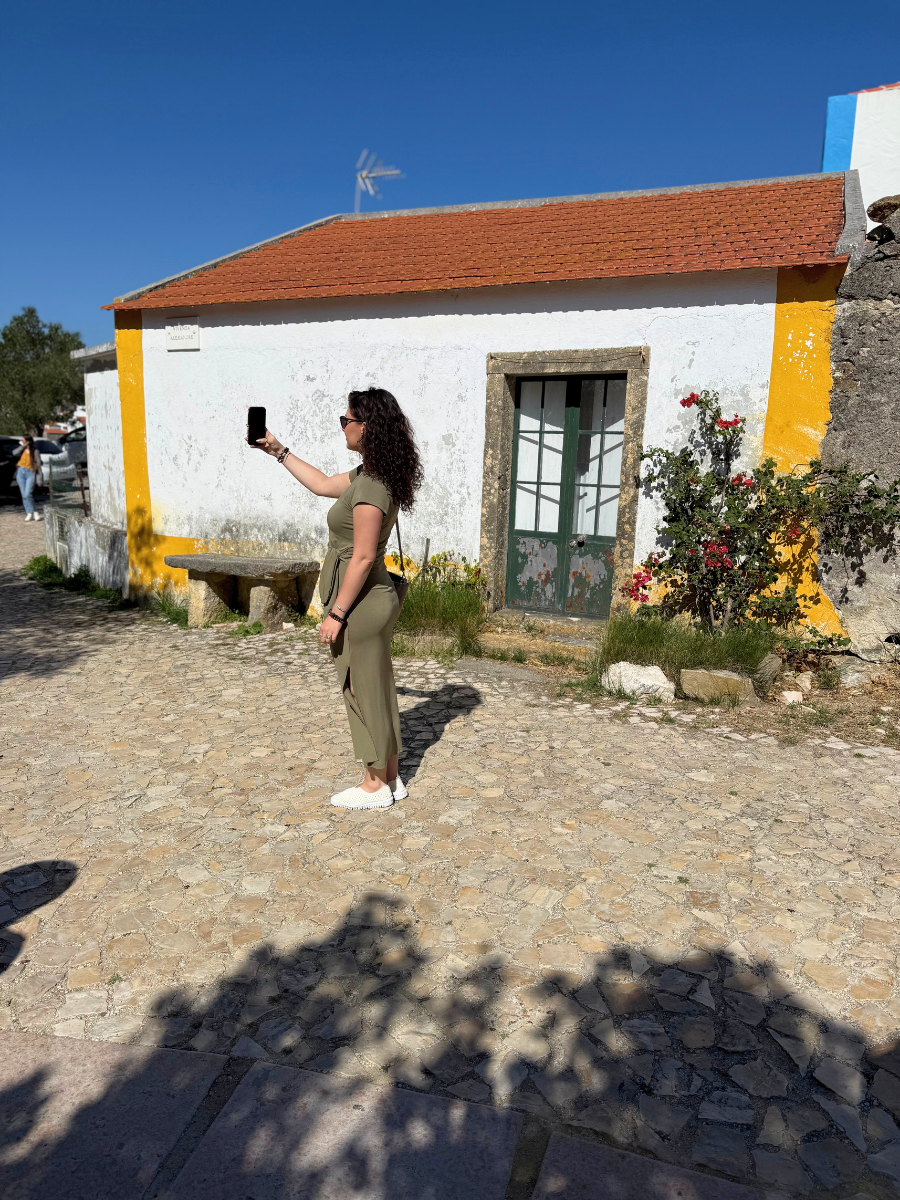 A woman in a green dress taking a photo on a cobblestone plaza next to a small, brightly painted, traditional Portuguese house with white, yellow, and orange accents.