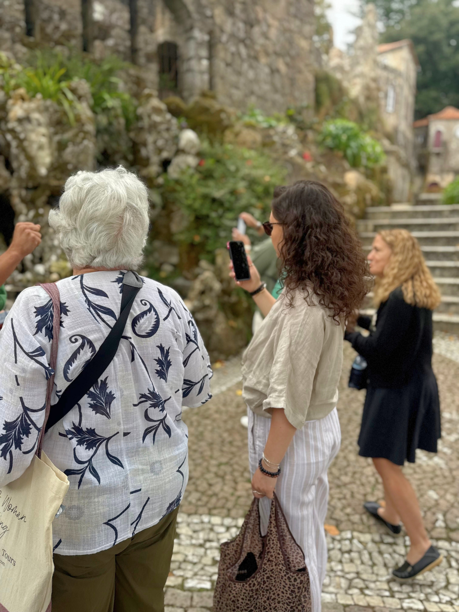 A group of three women standing on a cobblestone path near a mossy, rocky wall and stone staircase in the lush gardens of a historic site in Portugal.