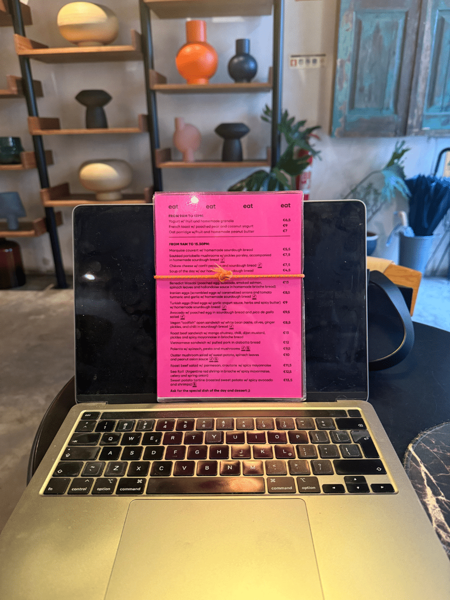 Photo of a laptop in Marquise cafe, with a bright pink menu displayed on the screen. Shelves in the background hold decorative pottery, adding a cozy and modern ambiance.