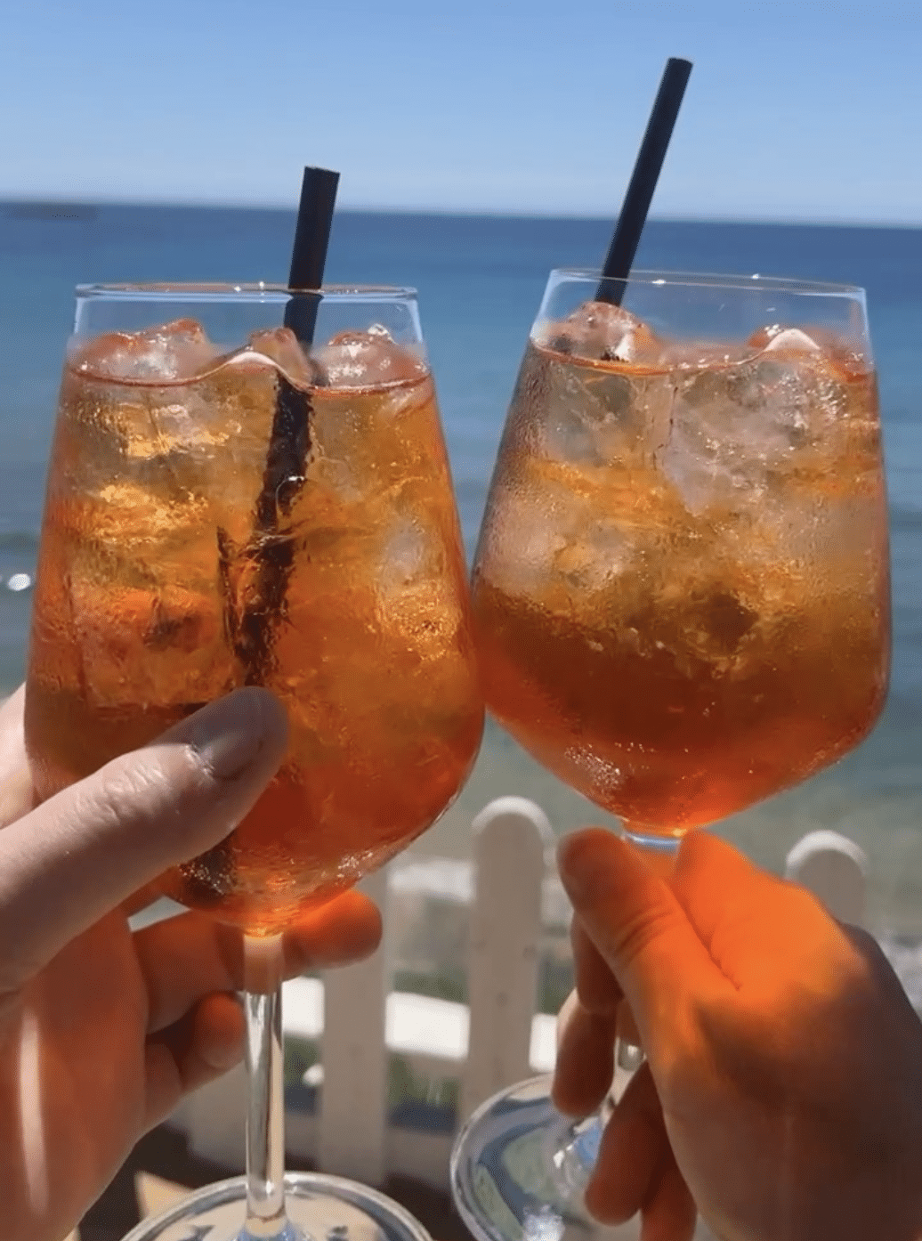 A close-up image of two Aperol Spritz drinks being held up in a toast. The drinks are in large wine glasses filled with ice, and the background shows a view of the sea and a clear blue sky.