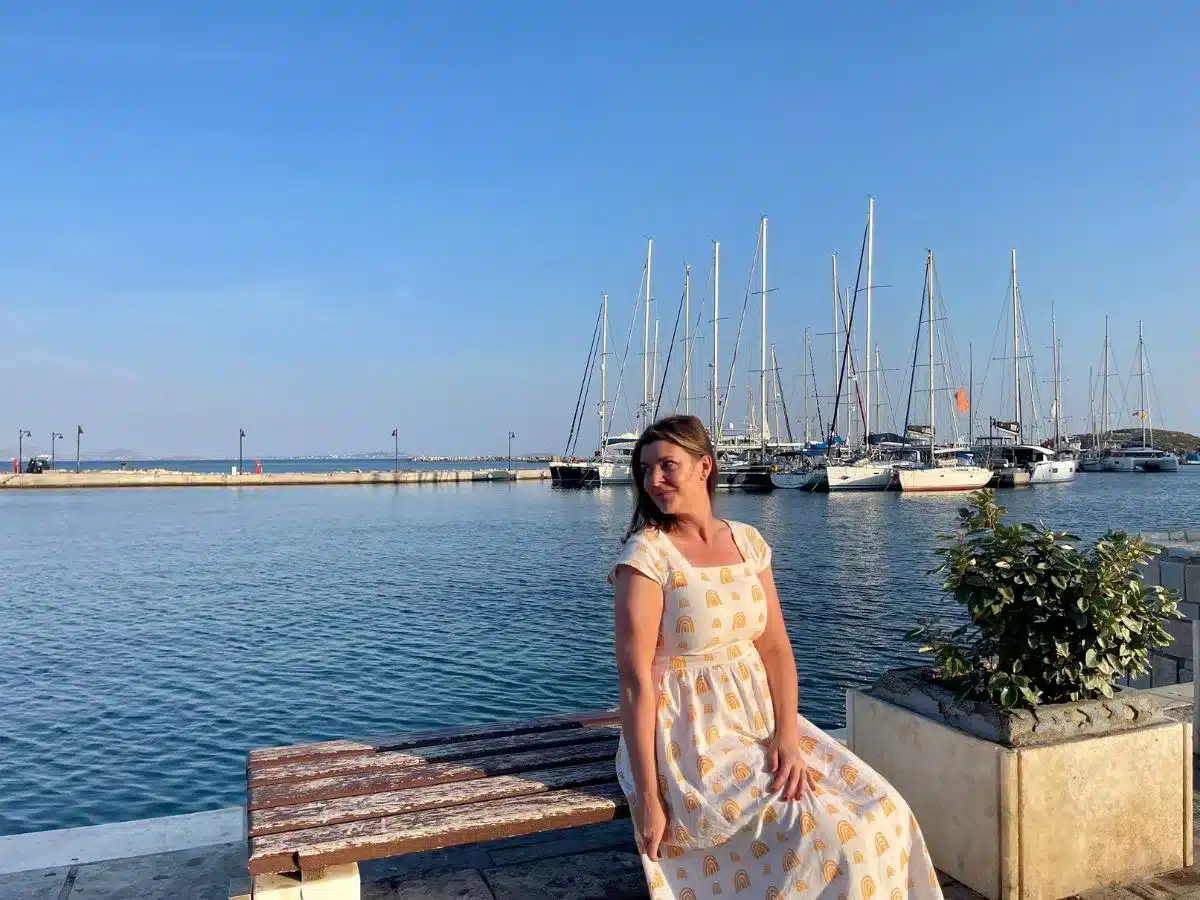 A picture of a woman sitting on the bench in the greek island of Naxos. The ocean and boats in the background. 