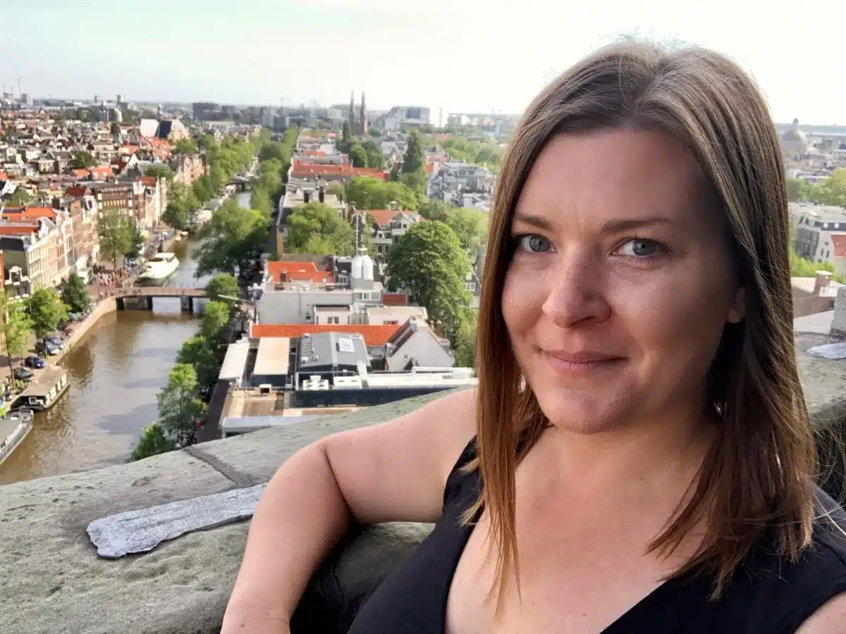A solo woman traveler smiles gently for a selfie with a panoramic view of a European city's classic architecture and serene canals in the background, suggesting she's enjoying her multi-city European adventure