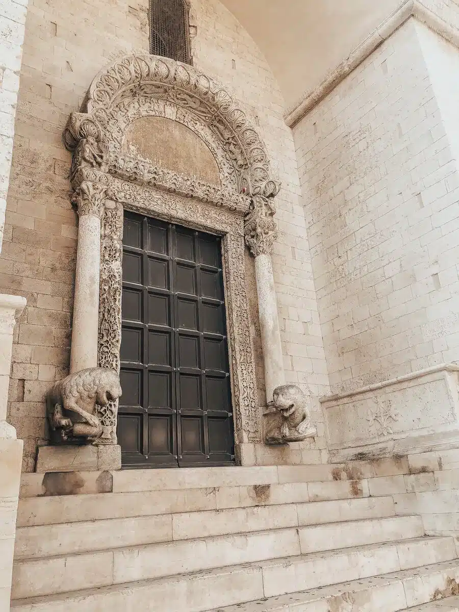 An ornate doorway of a church in Bari, Italy, featuring detailed stone carvings and statues of lions on either side of the steps. The door is large and dark, contrasting with the light stonework of the church.