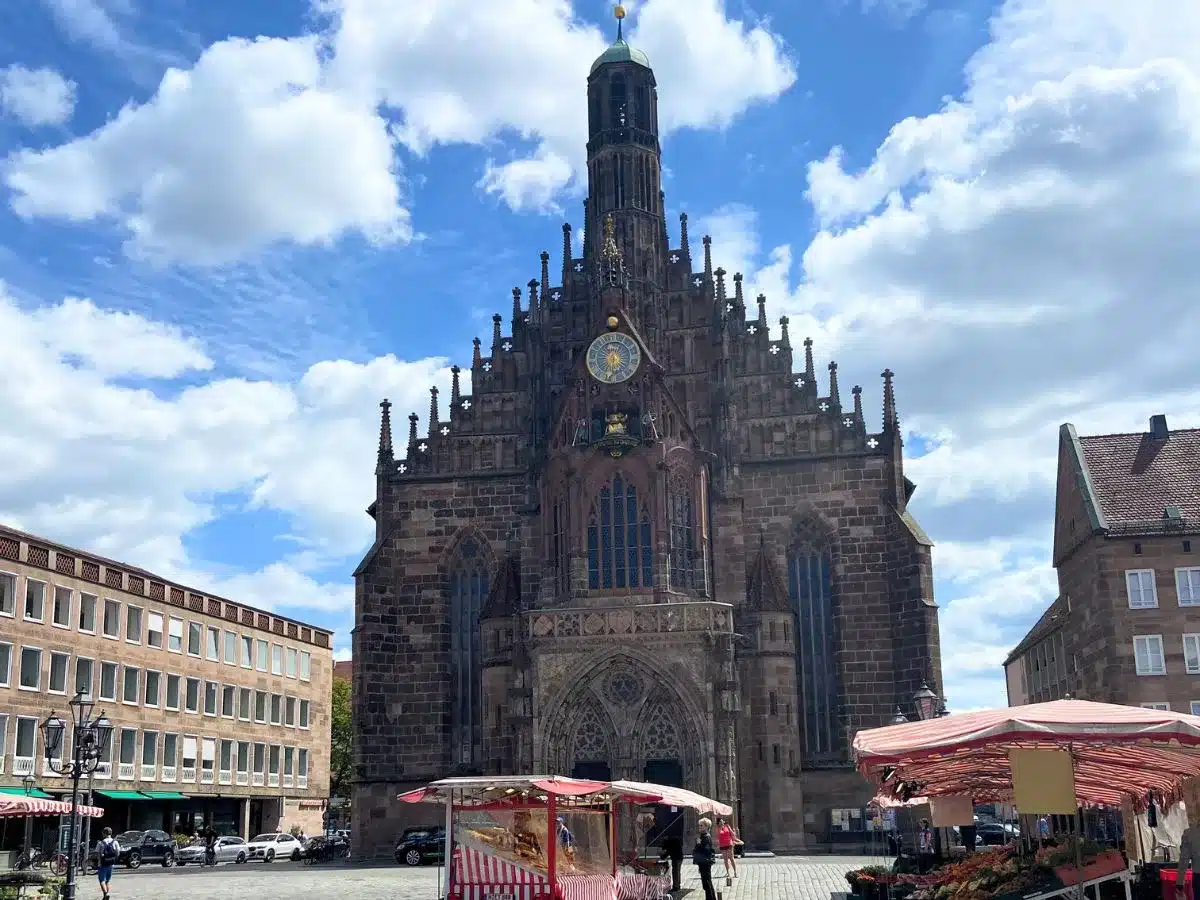The magnificent Gothic facade of The church of our lady under a sunny sky with fluffy clouds. The church's intricate stonework, grand spire, and ornate rose window are highlighted by the sun, showcasing this architectural marvel. In the foreground, market stalls and pedestrians create a lively scene in the spacious square.
